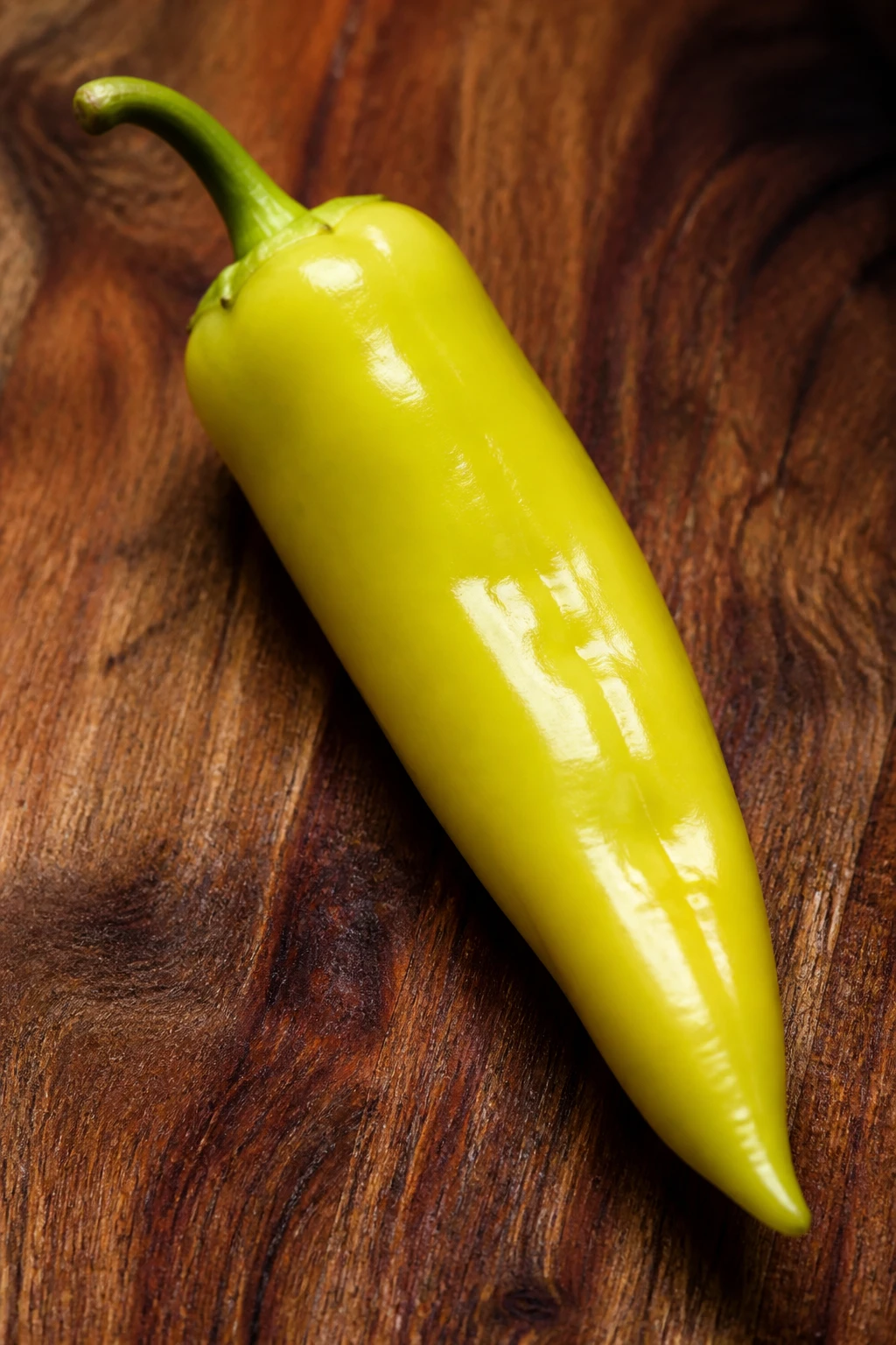 Closeup of a single glossy banana pepper resting on a wooden board, high contrast